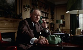 Movie still from “Goldfinger” (1964), directed by Guy Hamilton – An older man sitting at a desk talking on a phone; Medium shot, Low angle
