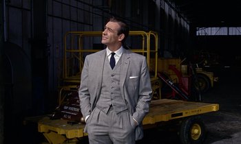 Movie still from “Goldfinger” (1964), directed by Guy Hamilton – A man in a suit and tie standing in front of a yellow cart; Medium shot, Low angle