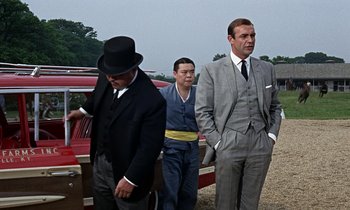 Movie still from “Goldfinger” (1964), directed by Guy Hamilton – A group of men standing next to each other near a red car; Medium shot, Low angle