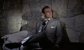Movie still from “Goldfinger” (1964), directed by Guy Hamilton – A man in a suit and tie sitting on a couch; Medium shot, Low angle
