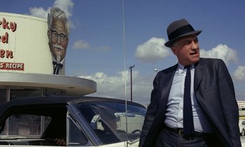 Movie still from “Goldfinger” (1964), directed by Guy Hamilton – A man in a suit and hat standing in front of a car; Medium shot, Low angle
