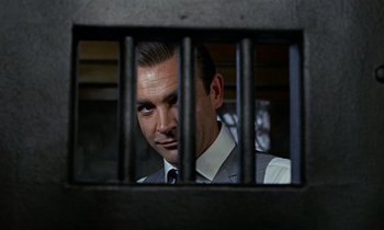 Movie still from “Goldfinger” (1964), directed by Guy Hamilton – A man wearing a suit and tie behind bars; Close Up shot, Low angle