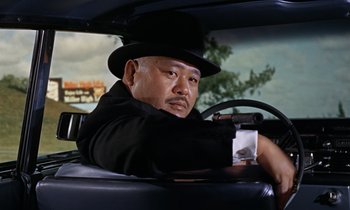 Movie still from “Goldfinger” (1964), directed by Guy Hamilton – A man sitting in the driver's seat of a car wearing a hat; Close Up shot, Low angle