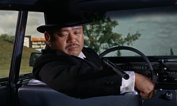 Movie still from “Goldfinger” (1964), directed by Guy Hamilton – A man in a top hat driving in a car; Medium shot, Low angle