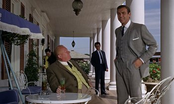 Movie still from “Goldfinger” (1964), directed by Guy Hamilton – A man in a suit and tie standing next to another man in a suit and tie; Medium shot, Low angle