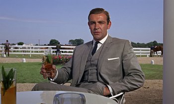 Movie still from “Goldfinger” (1964), directed by Guy Hamilton – A man in a suit and tie holding a glass of wine; Medium shot, Low angle