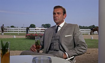 Movie still from “Goldfinger” (1964), directed by Guy Hamilton – A man in a suit and tie sitting at a table with a glass; Medium shot, Low angle