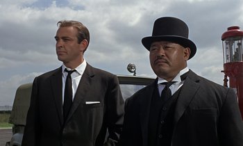 Movie still from “Goldfinger” (1964), directed by Guy Hamilton – Two men in suits and hats standing next to each other in front of a car; Medium shot, Low angle