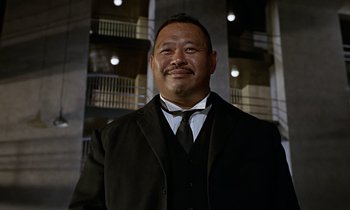 Movie still from “Goldfinger” (1964), directed by Guy Hamilton – A man in a suit and tie standing in front of a building; Close Up shot, Low angle