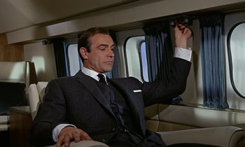 Movie still from “Goldfinger” (1964), directed by Guy Hamilton – A man in a suit and tie sitting in a car; Medium shot, Low angle