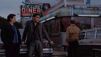 Movie still from “Goodfellas” (1990), directed by Martin Scorsese – A man in a suit and tie standing in front of a diner; Wide shot, Low angle