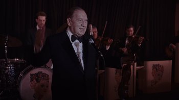 Movie still from “Goodfellas” (1990), directed by Martin Scorsese – An older man in a suit and bow tie singing into a microphone; Medium shot, Over the shoulder angle