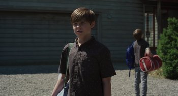 Movie still from “Goodnight Mommy” (2022), directed by Matt Sobel – A young boy standing in front of a building; Medium shot, Over the shoulder angle