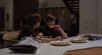 Movie still from “Goodnight Mommy” (2022), directed by Matt Sobel – Two young boys eating a piece of pizza at a dinner table; Medium shot, Over the shoulder angle