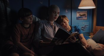 Movie still from “Goodnight Mommy” (2022), directed by Matt Sobel – A woman sitting next to two young boys reading a book; Medium shot, Over the shoulder angle