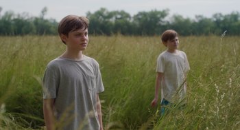 Movie still from “Goodnight Mommy” (2022), directed by Matt Sobel – Two young boys standing in a field of tall grass; Medium shot, Over the shoulder angle