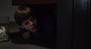 Movie still from “Goodnight Mommy” (2022), directed by Matt Sobel – A young boy is hiding under a cabinet; Close Up shot, High angle