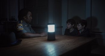 Movie still from “Goodnight Mommy” (2022), directed by Matt Sobel – Two boys and a woman sitting at a table with a lamp on top of it; Medium shot, Over the shoulder angle