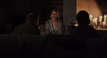 Movie still from “Goodnight Mommy” (2022), directed by Matt Sobel – A woman sitting in a chair in front of a group of people; Medium shot, Over the shoulder angle