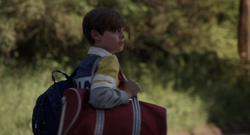 Movie still from “Goodnight Mommy” (2022), directed by Matt Sobel – A young boy is holding a bag of luggage; Medium shot, Over the shoulder angle