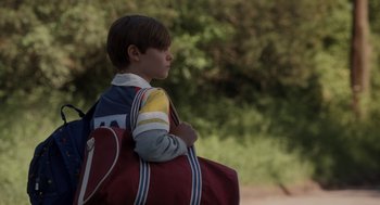 Movie still from “Goodnight Mommy” (2022), directed by Matt Sobel – A young boy with a red bag is standing outside; Medium shot, Over the shoulder angle