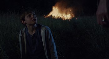 Movie still from “Goodnight Mommy” (2022), directed by Matt Sobel – A young boy sitting in the grass near a fire; Medium shot, Low angle