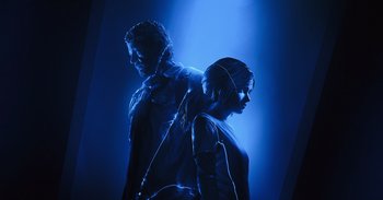 Movie still from “Gran Turismo” (2023), directed by Neill Blomkamp – A man and a woman standing next to each other in the dark; Medium shot, Overhead angle