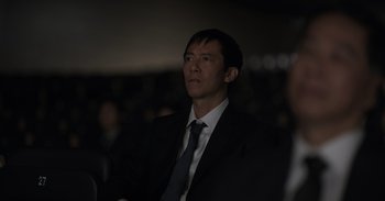 Movie still from “Gran Turismo” (2023), directed by Neill Blomkamp – A man wearing a suit and tie in a dark room; Close Up shot, Over the shoulder angle