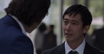 Movie still from “Gran Turismo” (2023), directed by Neill Blomkamp – A man in a business suit talking to another man in a business suit; Close Up shot, Over the shoulder angle