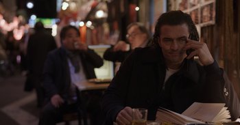 Movie still from “Gran Turismo” (2023), directed by Neill Blomkamp – A man sitting at a table with a glass of beer; Medium shot, Over the shoulder angle