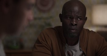 Movie still from “Gran Turismo” (2023), directed by Neill Blomkamp – A man sitting in front of a wall looking at the camera; Close Up shot, Over the shoulder angle