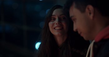 Movie still from “Gran Turismo” (2023), directed by Neill Blomkamp – A young woman smiles at the camera while a man looks on; Close Up shot, Over the shoulder angle