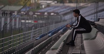Movie still from “Gran Turismo” (2023), directed by Neill Blomkamp – A man sitting on a bleacher in front of a fence; Wide shot, High angle