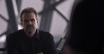 Movie still from “Gran Turismo” (2023), directed by Neill Blomkamp – A man sitting in front of another man in front of a window; Close Up shot, Over the shoulder angle