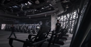Movie still from “Gran Turismo” (2023), directed by Neill Blomkamp – A man standing in front of a row of exercise machines; Extreme Wide shot, High angle