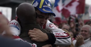 Movie still from “Gran Turismo” (2023), directed by Neill Blomkamp – A man hugging another man in front of a group of onlookers; Close Up shot, Over the shoulder angle