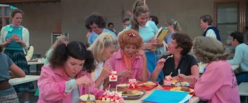 Movie still from “Grease” (1978), directed by Randal Kleiser – A group of people sitting at a table eating cake; Medium shot, Over the shoulder angle