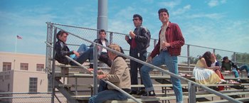 Movie still from “Grease” (1978), directed by Randal Kleiser – A group of men sitting on bleachers next to a chain link fence; Wide shot, Low angle