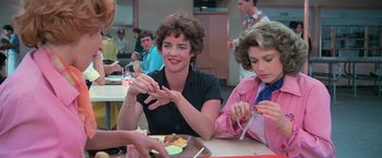 Movie still from “Grease” (1978), directed by Randal Kleiser – Two women sitting at a table with plates of food in front of them; Medium shot, Over the shoulder angle