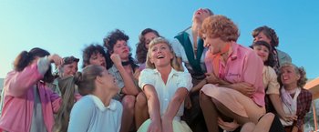 Movie still from “Grease” (1978), directed by Randal Kleiser – A group of people sitting around a woman laughing; Medium shot, Overhead angle