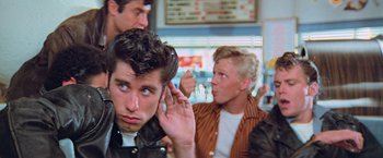 Movie still from “Grease” (1978), directed by Randal Kleiser – A young man with his hand up to his ear; Medium shot, Over the shoulder angle