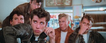 Movie still from “Grease” (1978), directed by Randal Kleiser – A group of young men standing next to each other in a restaurant; Medium shot, Low angle