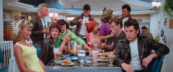 Movie still from “Grease” (1978), directed by Randal Kleiser – A group of people sitting at a table eating food; Wide shot, Over the shoulder angle