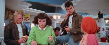 Movie still from “Grease” (1978), directed by Randal Kleiser – A man and a woman sitting in a room eating pizza; Medium shot, Over the shoulder angle