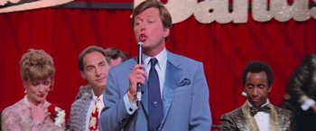 Movie still from “Grease” (1978), directed by Randal Kleiser – A man wearing a suit and tie holding a microphone; Close Up shot, Over the shoulder angle