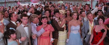 Movie still from “Grease” (1978), directed by Randal Kleiser – A group of people standing around each other in formal wear; Wide shot, Over the shoulder angle