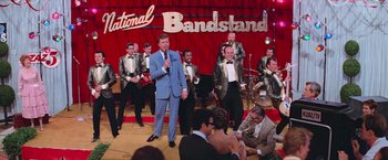 Movie still from “Grease” (1978), directed by Randal Kleiser – A man in a blue suit is on stage with a band; Wide shot, High angle
