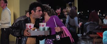 Movie still from “Grease” (1978), directed by Randal Kleiser – A man and a woman kissing while holding a box of food; Medium shot, Over the shoulder angle
