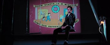 Movie still from “Grease” (1978), directed by Randal Kleiser – A man sitting on a swing in front of a mural; Wide shot, Low angle