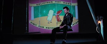 Movie still from “Grease” (1978), directed by Randal Kleiser – A man sitting on a swing in front of a screen; Wide shot, Low angle